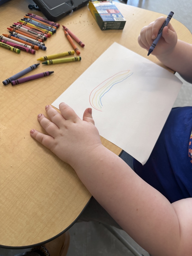 A child wearing a blue Sonic the Hedgehog t-shirt draws a rainbow on a white piece of paper with a blue crayon at a wooden table.