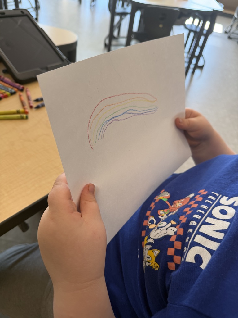A child in a blue Sonic the Hedgehog t-shirt holds up a hand-drawn crayon rainbow on a white piece of paper.