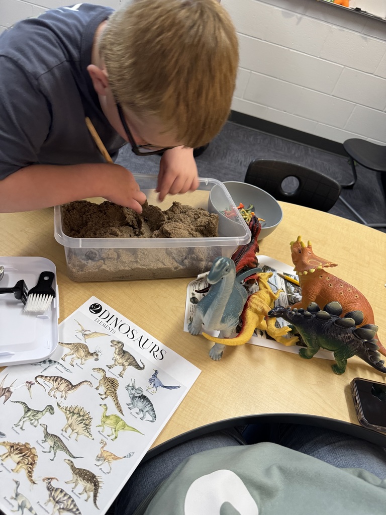 A boy with glasses leans over a table, focused on playing with kinetic sand and plastic dinosaur toys in a classroom.