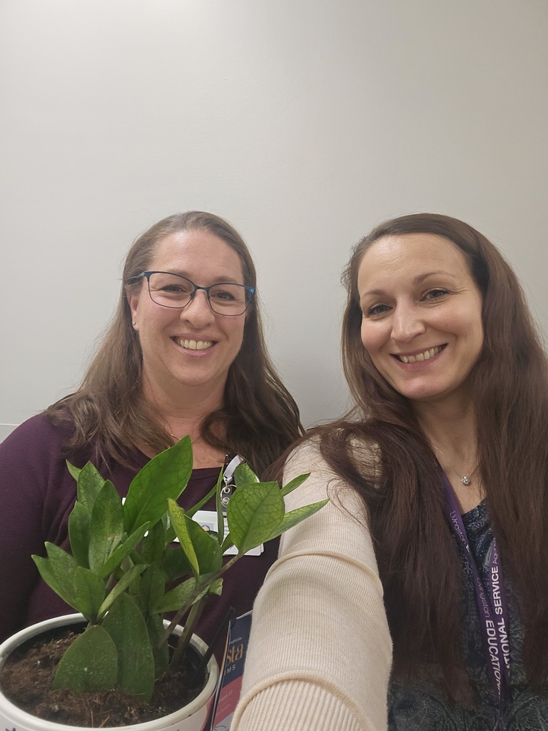 Two smiling women standing side-by-side in front of a plain, light-colored wall. The woman on the left, wearing glasses and a maroon top, holds a small potted plant with dark green, glossy leaves. The woman on the right, with long brown hair, wears a light-colored cardigan and a purple lanyard. Both women are looking toward the camera.