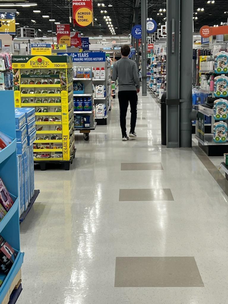 A man in a grey zip-up jacket and black track pants walks down a bright grocery store aisle, away from the camera. A large yellow display of Burpee seeds stands on the left.