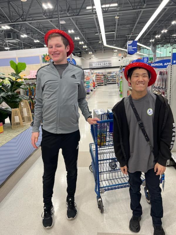 Two men stand together in a store aisle, smiling and wearing sparkly red, white, and blue top hats. One man wears a grey Adidas jacket, and the other wears a black hoodie and a grey t-shirt.