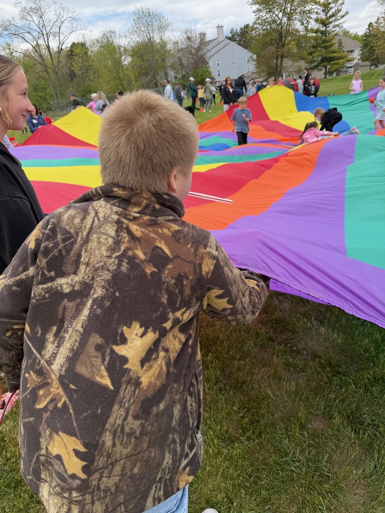 A young boy wearing a camo jacket is holding the edge of a giant parachute, lifting it up high. Kids can be seen running around in the background, walking on top of the multi-colored parachute.