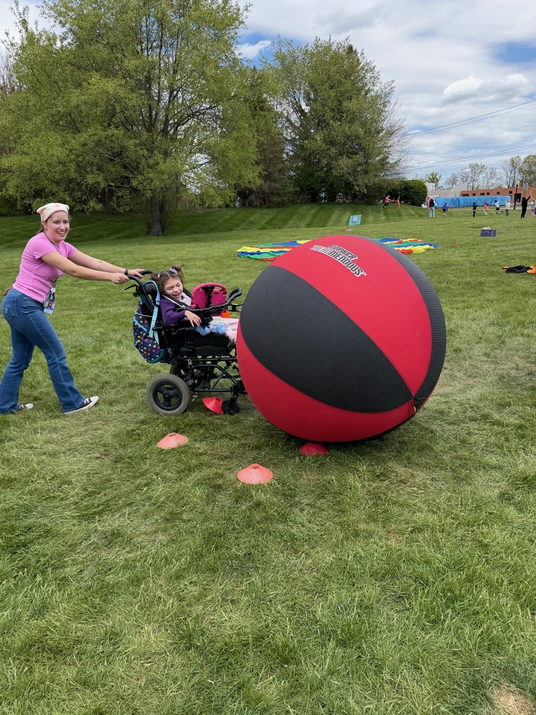 A woman pushes a young girl in a wheelchair while the young girl is pushing a giant ball on a grassy field. 