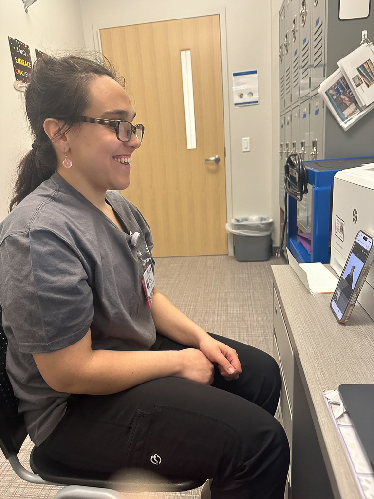 A person with dark hair pulled back and wearing glasses and grey scrubs sits in a chair, smiling while looking at a smartphone propped up on a desk. The phone appears to be displaying a video call or a video. The person is in a room with a closed wooden door, grey lockers, and a printer on the desk.