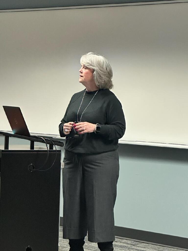A woman with silver hair standing at a podium next to a laptop, giving a presentation in front of a white screen
