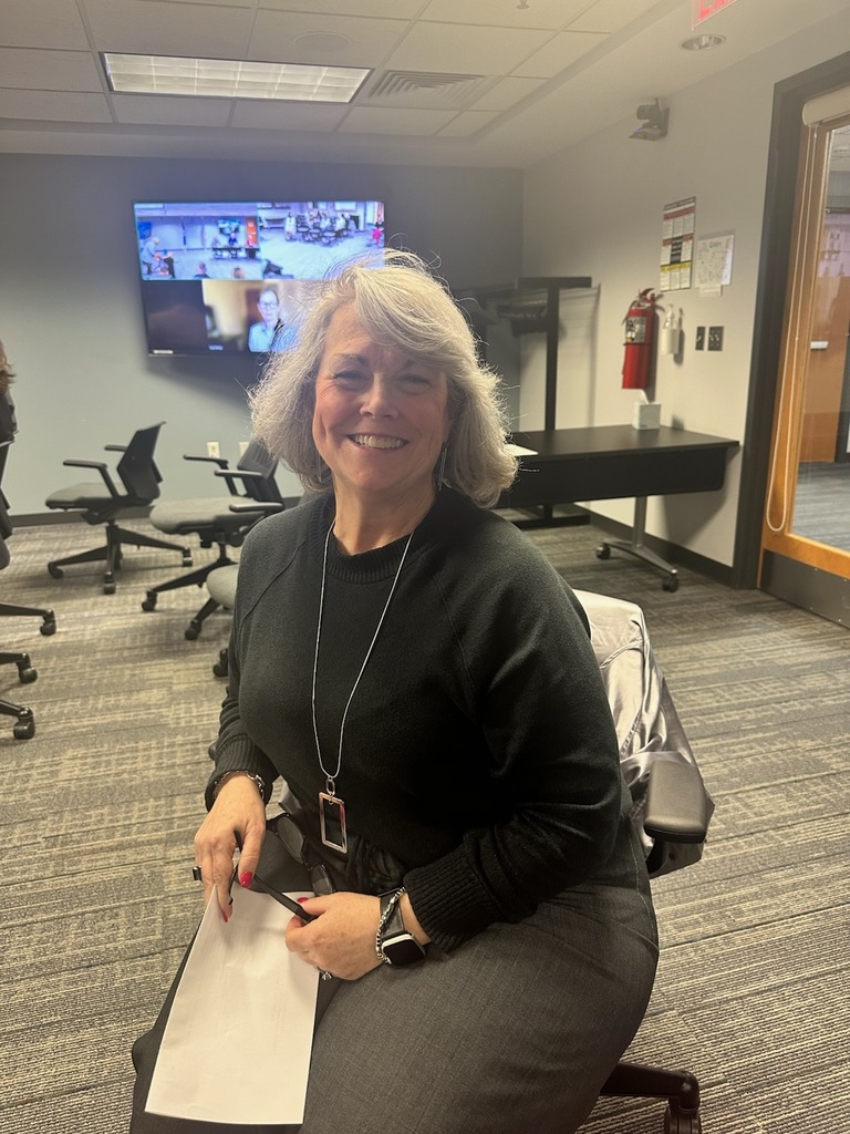 A smiling woman with silver hair sitting in a professional office or conference room, wearing a black sweater and a long silver pendant necklace.