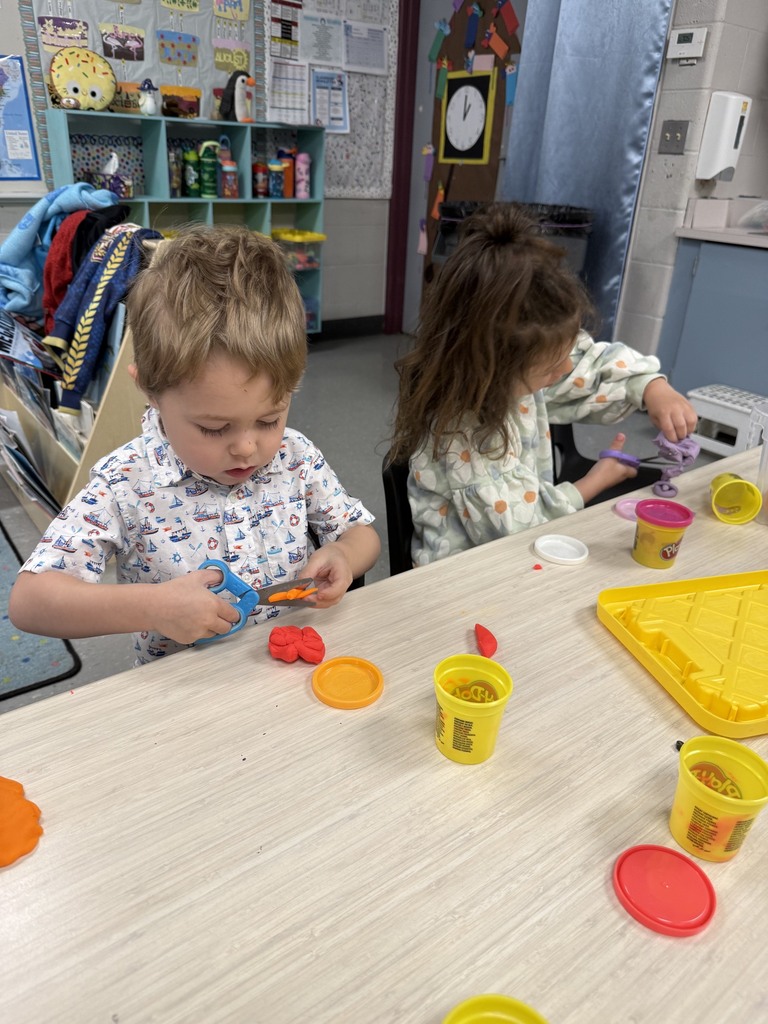 Two preschoolers sit at a classroom table using scissors and playdough to strengthen fine motor skills and practice hand coordination.