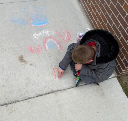 A young boy in a grey winter parka kneels on a sidewalk, focused on creating colorful chalk drawings like hearts and rainbows next to a brick wall.