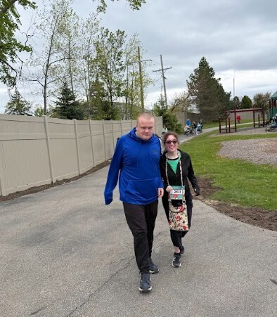 A young woman walks with a floral purse around her neck next to a young man in a blue hoodie and black pants while they both smile at the camera.