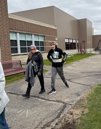 A young man dressed in a black hoodie and grey pants has his hands on his hips as he walks next to a female staff member