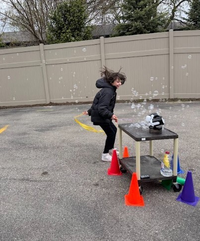 A young girl in a black coat and black pants is jumping while playing with bubbles