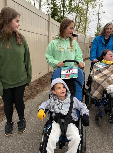 A boy in a stroller smiles at the camera while being pushed by a young girl on an outdoor track.