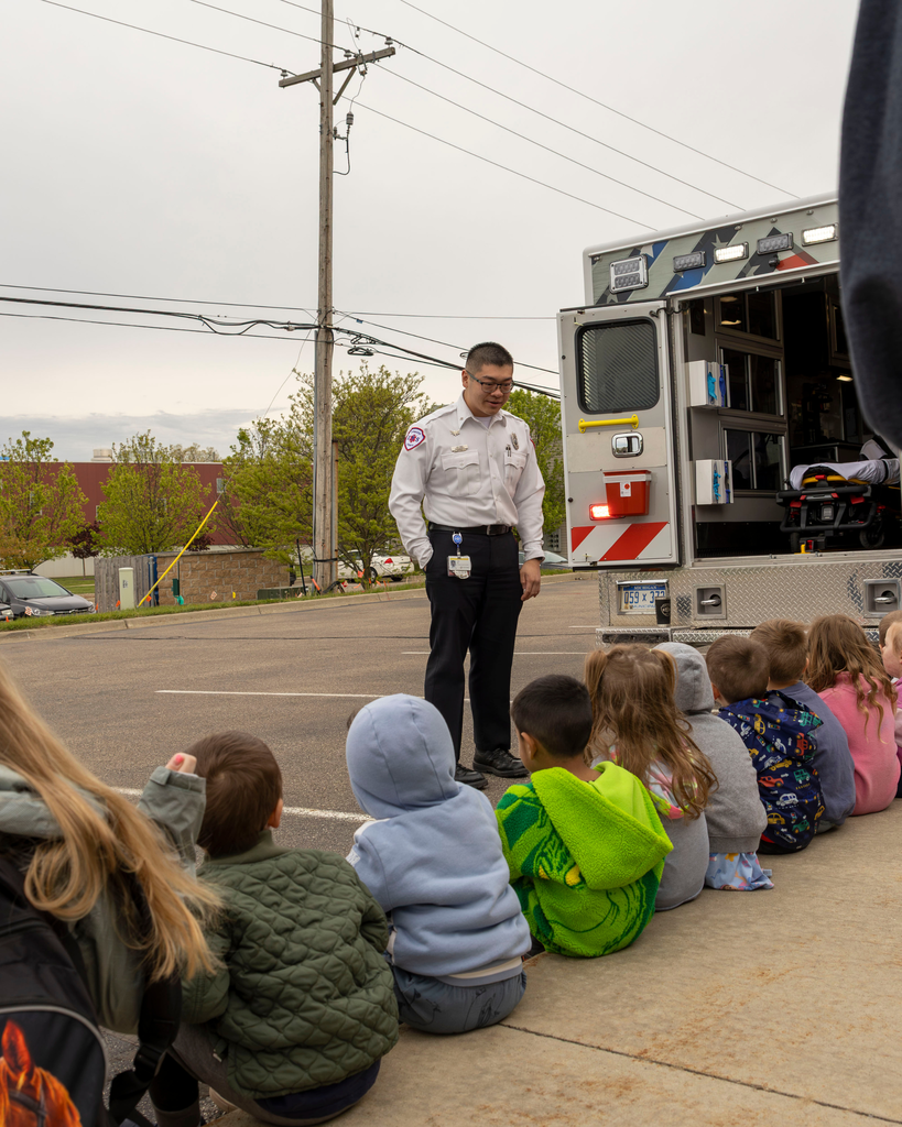 A wide outdoor shot of a male paramedic in a white uniform shirt and dark pants standing behind an open ambulance door. He is speaking to a line of young children who are sitting on the curb of a sidewalk, listening attentively. The scene takes place in a parking lot on an overcast day with trees and power lines in the background.
