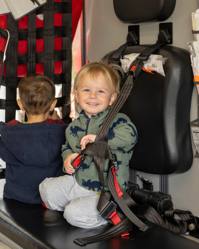 A close-up shot inside an ambulance showing a smiling toddler with blonde hair sitting on a black bench seat. The toddler is wearing a green fleece jacket with a dinosaur pattern and grey sweatpants, holding onto a black safety harness strap. In the background, another young child in a dark blue hoodie is sitting with their back to the camera, next to a red equipment wall with black netting.