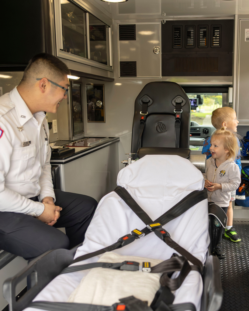 An interior view of an ambulance where a male paramedic sits on the bench seat, smiling at two young children standing near the stretcher. The stretcher is in the foreground, covered in white sheets with black safety straps laid across it. The ambulance interior features storage cabinets and a large black medical chair in the background.