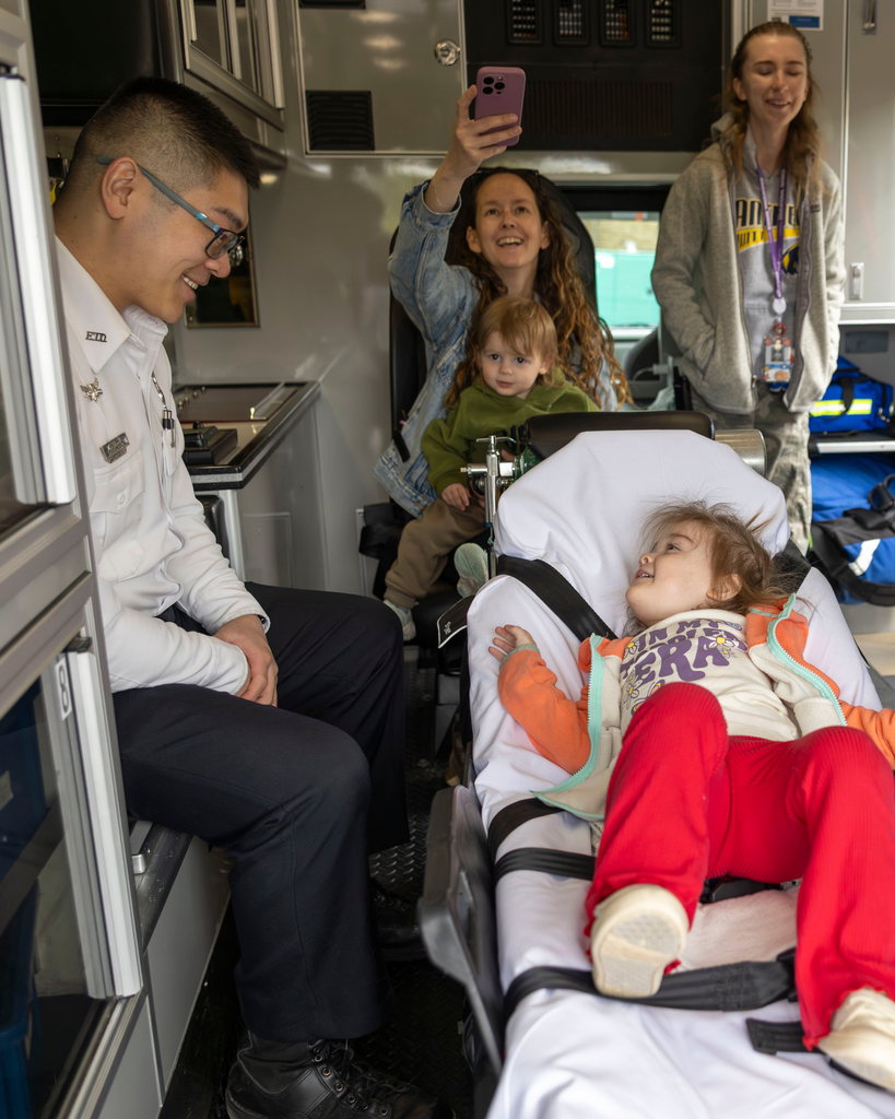 A medium shot inside an ambulance showing a male paramedic smiling down at a young girl lying on a stretcher. The girl, wearing red pants, a white shirt, and an orange jacket, looks up happily at him. In the background, a woman holds a toddler and takes a selfie with her phone, while another young woman stands nearby smiling.