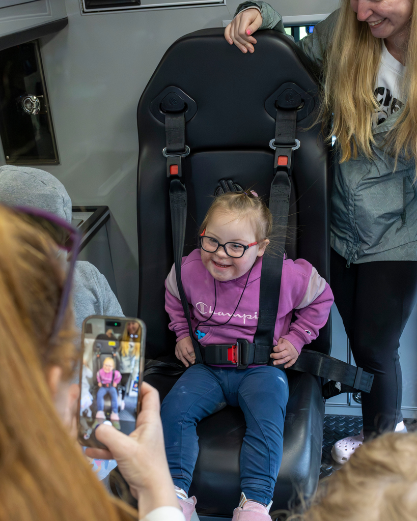 A close-up shot inside an ambulance showing a young girl with glasses and Down syndrome smiling joyfully while buckled into a black medical harness seat. An adult woman stands to her right, and in the foreground, another person holds up a smartphone to take a photo of the smiling girl.