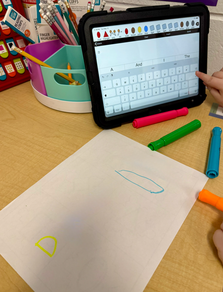A high-angle shot shows a child using an educational writing app on a tablet next to a piece of paper with a hand-drawn yellow letter "D," surrounded by colorful markers and classroom supplies on a wooden desk.