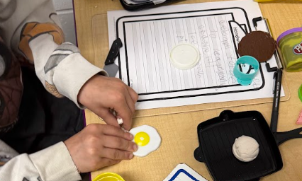 Close-up of a child's hands shaping white and yellow playdough into a toy fried egg next to a handwriting worksheet on a clipboard.