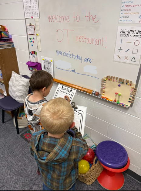 Two young boys hold clipboards and read a whiteboard that says "Welcome to the OT restaurant!" in an occupational therapy room setting.