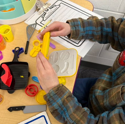 Over-the-shoulder view of a child using a yellow playdough mold at a desk scattered with toy kitchen utensils and handwriting practice sheets.