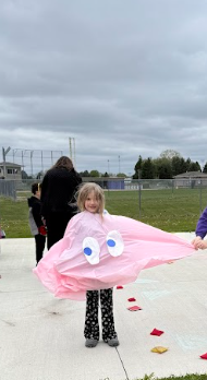 A smiling young girl stands outdoors on a concrete path while wearing a large, homemade pink Pac-Man ghost costume.