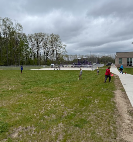 A group of children spread out across a large green lawn, playing outdoor games near a fenced-in school playground under a cloudy sky.