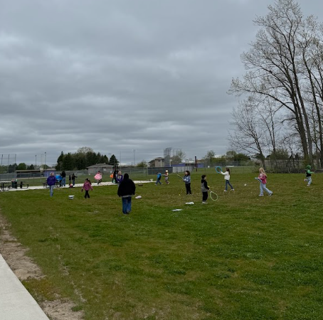 Several school children participate in various field day like activities, including throwing frisbees and running, on an open grassy field.