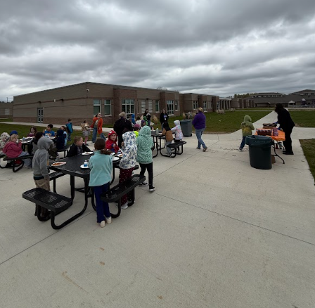 A large group of elementary school students and staff gather at outdoor picnic tables next to a brick building for a lunchtime pizza party.