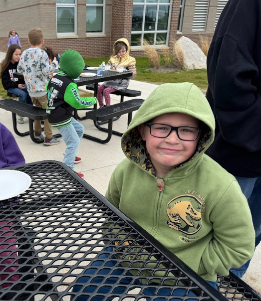 A young boy wearing glasses and a green Jurassic World hooded sweatshirt smiles warmly while sitting at an outdoor picnic table with classmates.