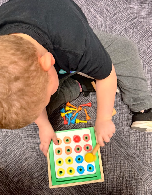 A young boy sitting on a carpeted floor uses a small wooden mallet to pound colorful plastic pegs into a matching grid toy.