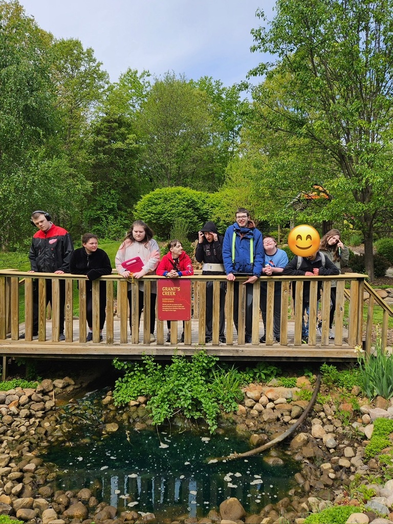Nine students stand on a wooden bridge over a small pond with trees in the background