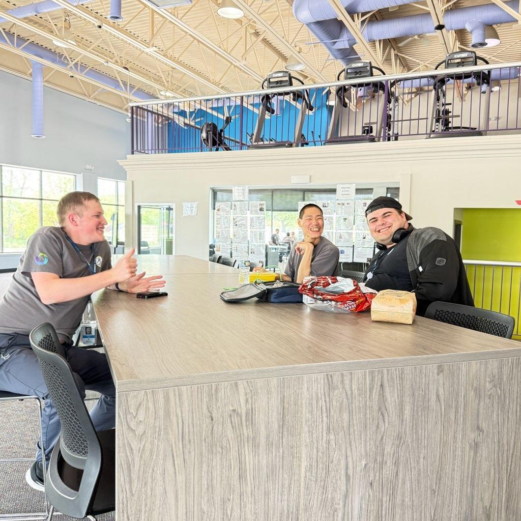 A group of young men sit at a counter during a meal break, viewed from a slight distance. A mezzanine balcony with fitness equipment overlooks the seating area.