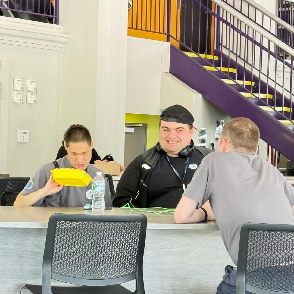 Three young men are seated at a high-top counter, talking and smiling. A purple and yellow staircase is visible in the background.