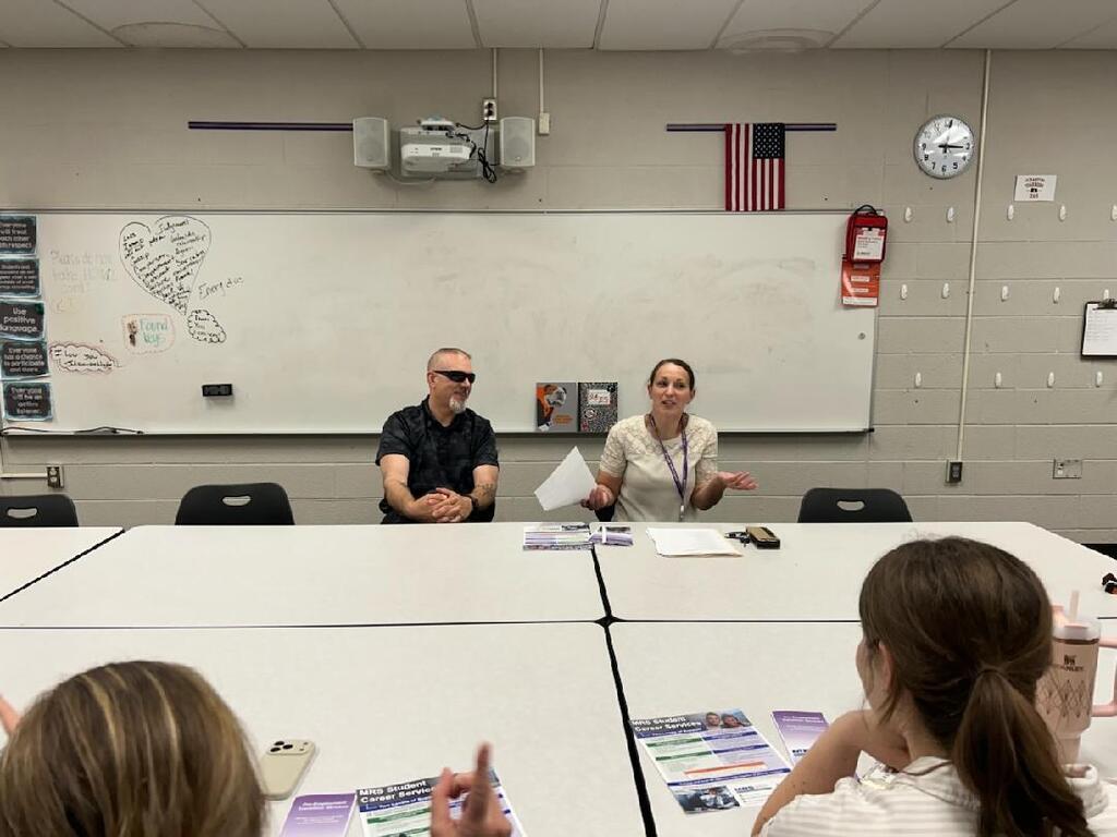 A man and a woman sit at a large white table at the front of a classroom, facing two attendees in the foreground whose backs are partially turned to the camera. A large whiteboard is behind the speakers.