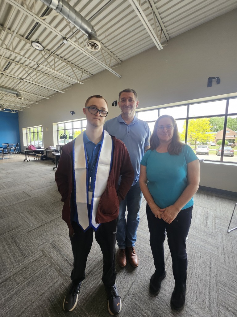 A young man wearing a graduation stole stands between an older man and woman inside a spacious room with large windows and high ceilings.