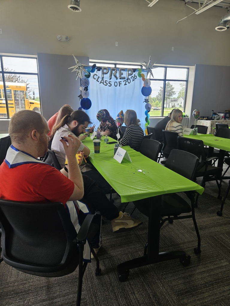 People sit along a long table covered with a bright green tablecloth. In the background, a light blue backdrop is decorated with balloons, stars, and hanging decorations that read "PREP CLASS OF 2026." A yellow school bus is visible through the window.