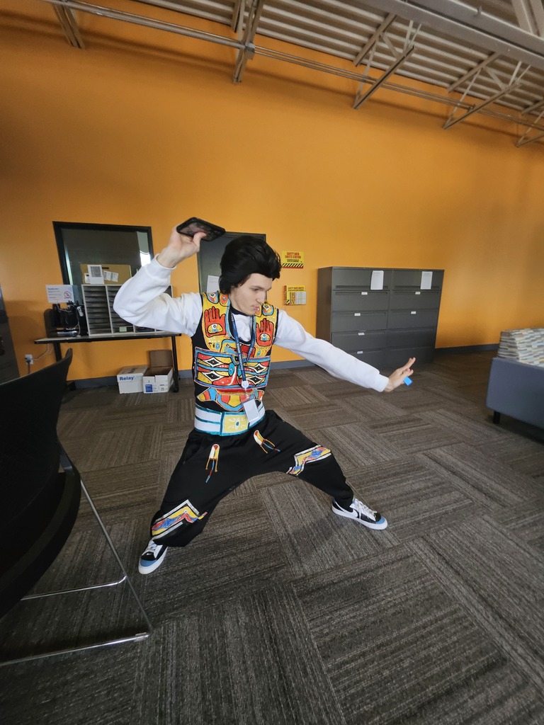 A young man with black hair strikes a dynamic, wide-legged action pose against a bright orange wall. He is wearing a white sweatshirt under a colorful, pattern-filled vest and matching black pants with geometric designs