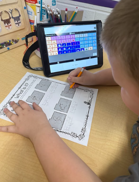 A young boy writes on a school worksheet at a desk with a tablet displaying an augmentative and alternative communication (AAC) grid propped up in front of him.
