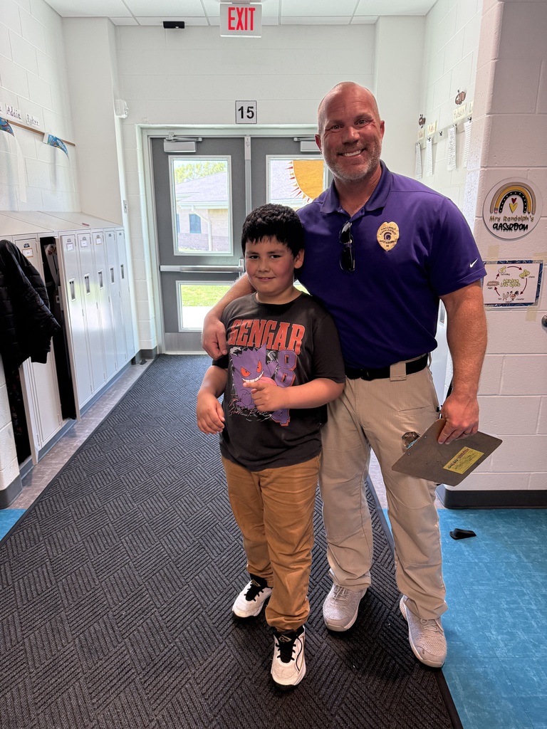 A smiling school officer in a purple polo shirt stands with his arm around a young student in a Pokémon t-shirt inside a school hallway.