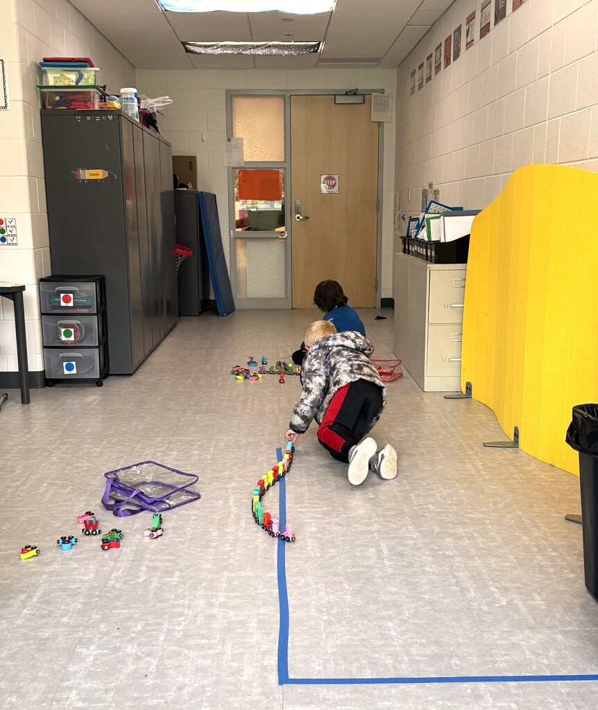 Two children play on a school hallway floor, with one child lining up small colorful toy trains along a blue tape line and the other boy sitting surrounded by toy trains.