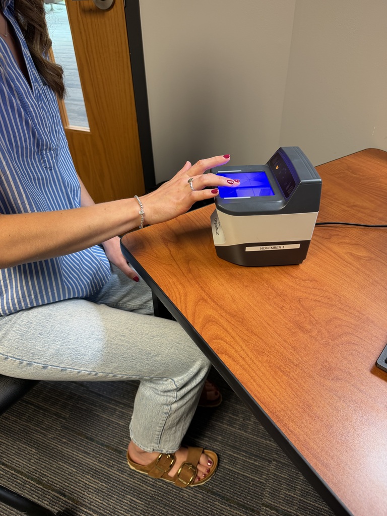 A woman sits at a wooden desk and places her right hand onto the blue-lit glass surface of a Crossmatch biometric fingerprint scanner for a background check or identity verification.