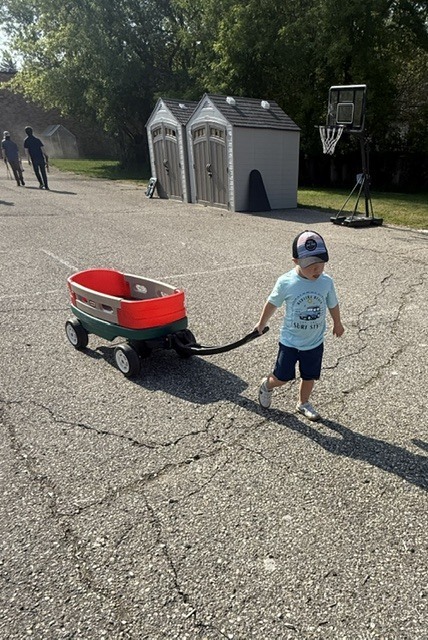 A preschooler wearing a hat pulls a red wagon across a playground blacktop area on a sunny day. Storage sheds and a basketball hoop are visible in the background.
