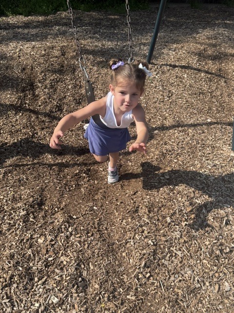 A preschooler with pigtails swings independently on a playground swing over wood chips while reaching forward with a smile.