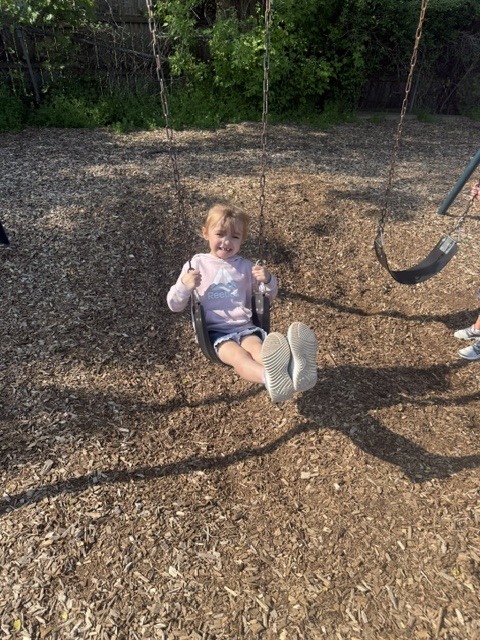 A smiling preschooler in a pink sweatshirt swings on a playground swing outdoors over wood chips on a sunny day.