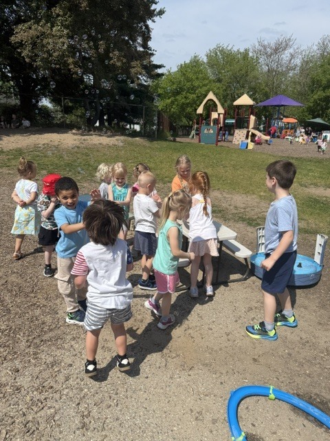 A group of preschoolers play outside during Bubble Day, reaching for bubbles and gathering around playground equipment on a sunny day.