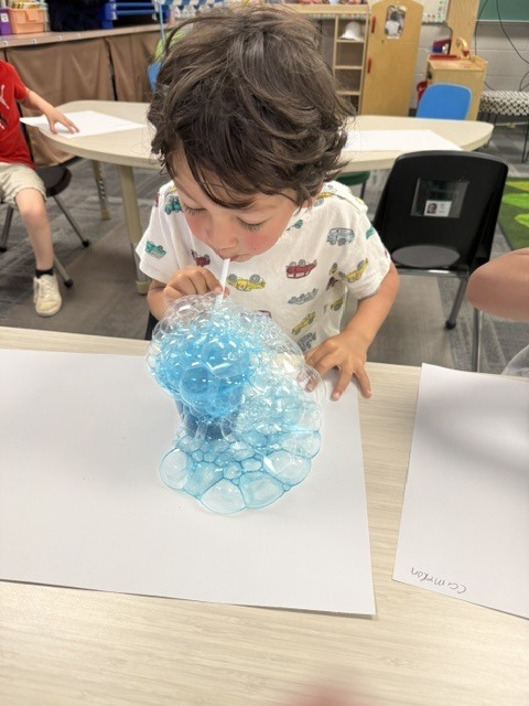 A preschooler blows through a straw into a blue bubble mixture, creating a large mound of bubbles on white paper at a classroom table.