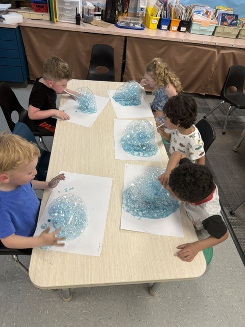 Preschoolers gathered around a table create overflowing blue bubbles with straws and cups during a hands-on science activity.