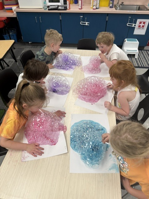 A group of preschoolers sit around tables blowing bubbles through straws into colorful soapy mixtures, creating large bubble formations on paper during a science activity.